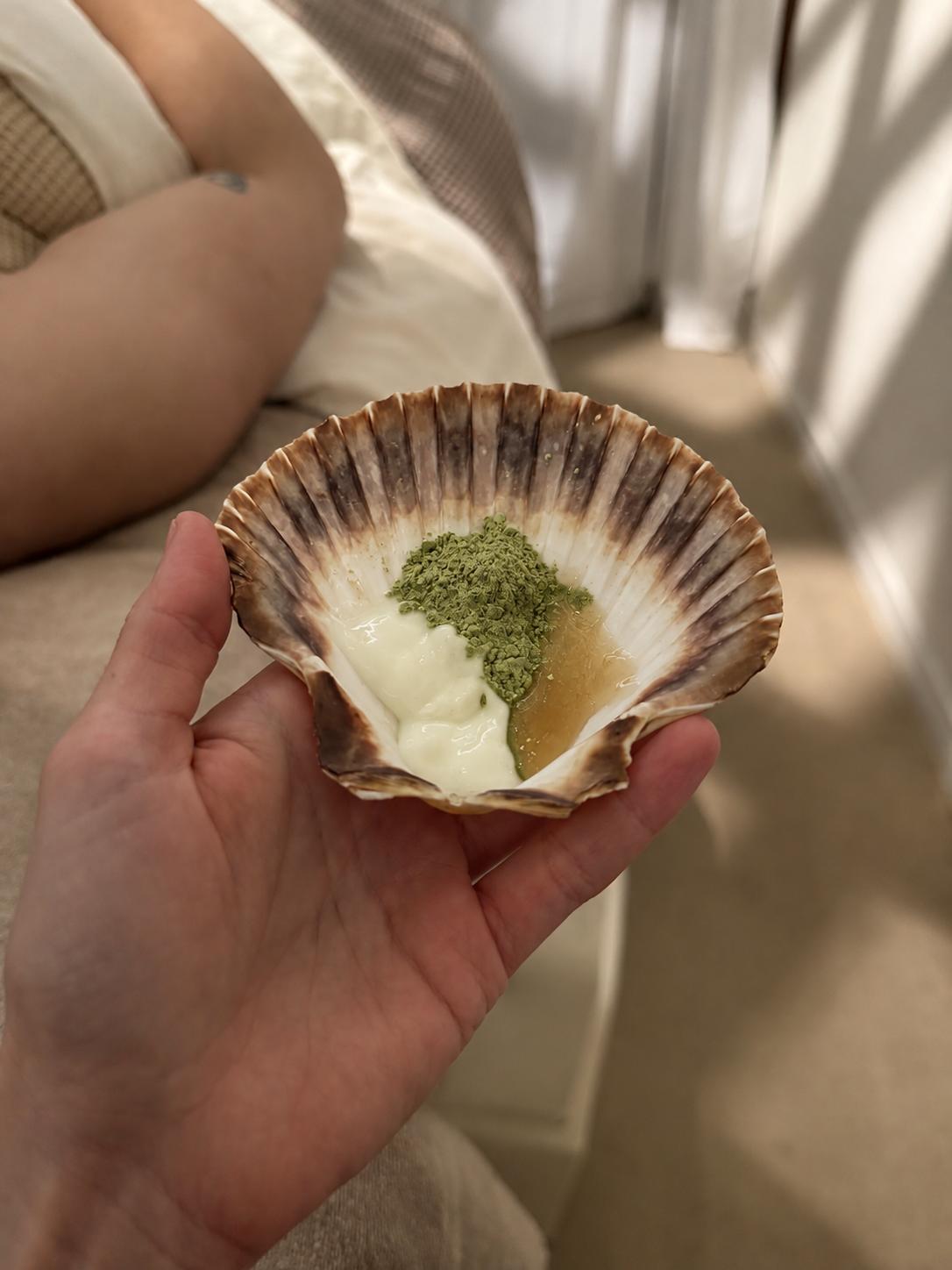 Mano sosteniendo una concha con una mezcla fresca de matcha y otros ingredientes para preparar un tratamiento personalizado